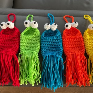 May include: Five colorful, handmade crochet monster pouches. Each pouch is a different color: red, green, blue, orange, and yellow. They have large, googly eyes and fringe at the bottom. Each pouch has a loop at the top for hanging.