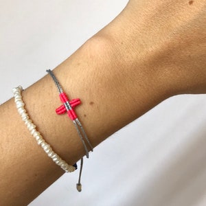 Può includere: Un braccialetto di corda grigio e rosa con un piccolo ciondolo a forma di croce. Il braccialetto è legato con un nodo e ha una perla argentata all'estremità.
