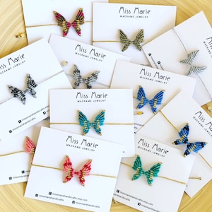 Op de afbeelding: Assortiment van kleurrijke macramé vlinderarmbanden met gouden accenten. Elke armband wordt getoond op een witte kaart met de tekst "Miss Marie Macrame Jewelry".