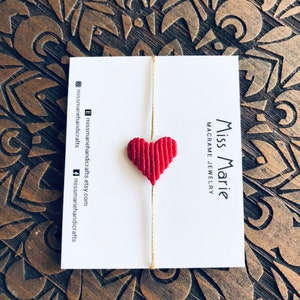 Puede incluir: Llavero de macramé en forma de corazón rojo que cuelga de una cuerda dorada. El llavero está en una tarjeta blanca con el texto "Miss Marie Macramé Joyería".