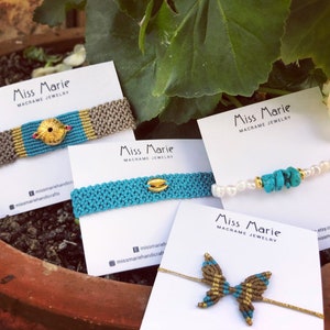 May include: Four macrame bracelets with different charms. The bracelets are displayed on white cards with the text "Miss Marie Macrame Jewelry". The charms include a gold disc with a turquoise centre, a gold shell, a cluster of turquoise stones and pearls, and a gold butterfly.