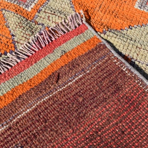 K&ouml;nnte beinhalten: Nahaufnahme eines bunten gewebten Teppichs mit einem geometrischen Muster in Orange-, Rot-, Braun- und Gr&uuml;nt&ouml;nen. Der Teppich hat einen Fransenrand und besteht aus einem dicken, strukturierten Material.
