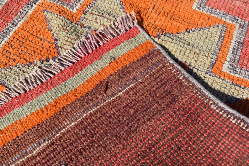 K&ouml;nnte beinhalten: Nahaufnahme eines bunten gewebten Teppichs mit einem geometrischen Muster in Orange-, Rot-, Braun- und Gr&uuml;nt&ouml;nen. Der Teppich hat einen Fransenrand und besteht aus einem dicken, strukturierten Material.