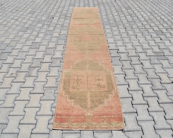 Puede incluir: Una alfombra de pasillo rectangular de estilo vintage con un aspecto descolorido. La alfombra presenta un diseño geométrico central en verde oliva y un borde salmón. La alfombra está colocada sobre una superficie gris estampada. Mide unos 200 cm de largo.