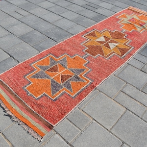 K&ouml;nnte beinhalten: Ein roter, orangefarbener und brauner Teppich mit geometrischem Muster. Der Teppich liegt auf einer grauen Betonfl&auml;che.