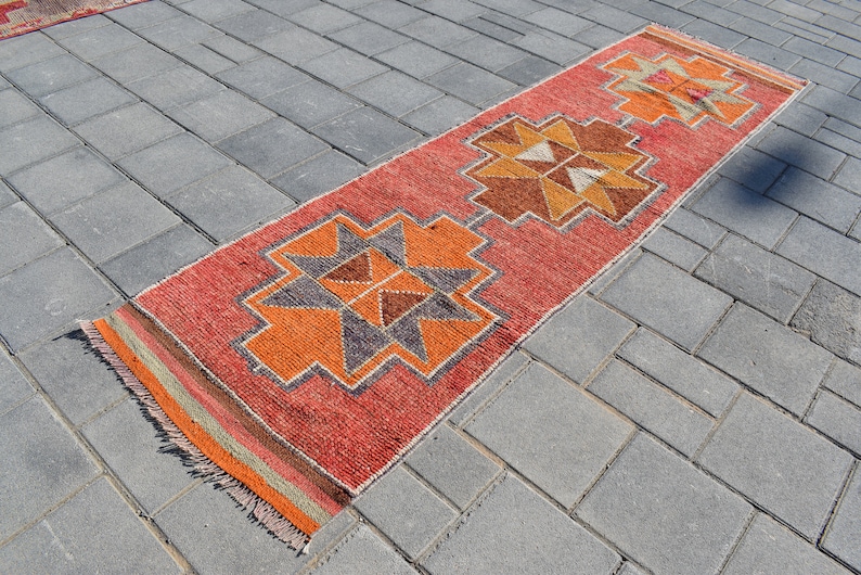 K&ouml;nnte beinhalten: Ein roter, orangefarbener und brauner Teppich mit geometrischem Muster. Der Teppich liegt auf einer grauen Betonfl&auml;che.
