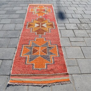 K&ouml;nnte beinhalten: Ein langer, rechteckiger Teppich mit einem geometrischen Muster in Orange-, Rot-, Braun- und Graut&ouml;nen. Der Teppich hat an beiden Enden Fransen.