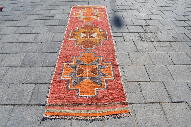 K&ouml;nnte beinhalten: Ein langer, rechteckiger Teppich mit einem geometrischen Muster in Orange-, Rot-, Braun- und Graut&ouml;nen. Der Teppich hat an beiden Enden Fransen.