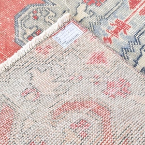May include: Close-up of a beige rug with a red and blue geometric pattern. The rug has a fringe border and a white tag with the text "No. 671 111.00 HAND MADE IN TURKEY".