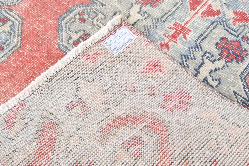 May include: Close-up of a beige rug with a red and blue geometric pattern. The rug has a fringe border and a white tag with the text "No. 671 111.00 HAND MADE IN TURKEY".