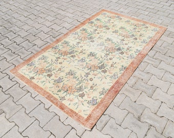 May include: A vintage floral patterned rug with a cream background and a peach-colored border. The rug has a distressed look and is laid out on a gray brick surface.