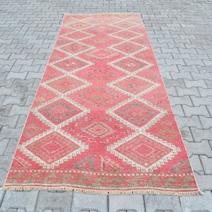 Op de afbeelding: Een roze en crèmekleurig tapijt met een ruitpatroon. Het tapijt heeft een franje rand en is gemaakt van wol.