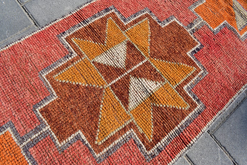 K&ouml;nnte beinhalten: Nahaufnahme eines roten, orangefarbenen und braunen Teppichs mit geometrischem Muster. Der Teppich hat einen Fransenrand und ist aus gewebten Fasern gefertigt.
