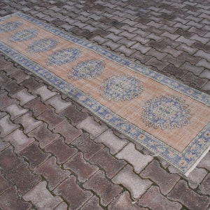 Könnte beinhalten: Ein langer, rechteckiger Teppich mit einem hellpfirsichfarbenen Hintergrund und einem blauen und weißen Blumenmuster. Der Teppich hat einen Fransenrand.