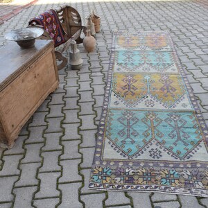 Könnte beinhalten: Ein langer, rechteckiger Teppich mit einem farbenfrohen geometrischen Muster in Blautönen, Gelb und Braun. Der Teppich ist aus gewebtem Stoff gefertigt und hat einen Fransenrand. Der Teppich liegt auf einer grauen Steinplatte.