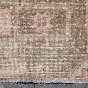 May include: A vintage rug with a brown and pink geometric pattern. The rug has a fringe border and is made of a woven material.