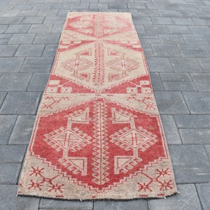 Op de afbeelding: Een rood en beige loper met een geometrisch patroon. De loper heeft een verweerde uitstraling en is gemaakt van wol.