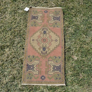 Peut inclure: Un tapis rectangulaire tissé à la main avec un motif central en losange et des motifs géométriques dans les tons de rose, de vert et de bleu marine. Le tapis mesure environ 30 cm de large et 61 cm de long, avec une bordure à franges.