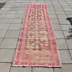 Könnte beinhalten: Ein langer, schmaler Teppich mit einem geometrischen Muster in Rosa und Braun. Der Teppich hat an beiden Enden Fransen.