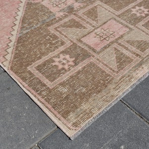 May include: A vintage rug with a geometric pattern in shades of pink and brown. The rug has a fringed edge and is made of wool.