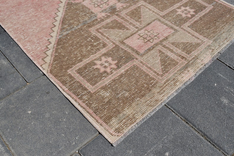 May include: A vintage rug with a geometric pattern in shades of pink and brown. The rug has a fringed edge and is made of wool.