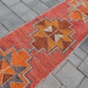 K&ouml;nnte beinhalten: Ein roter, orangefarbener und brauner Teppich mit geometrischem Muster. Der Teppich liegt auf einer grauen Betonfl&auml;che.