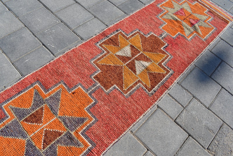 K&ouml;nnte beinhalten: Ein roter, orangefarbener und brauner Teppich mit geometrischem Muster. Der Teppich liegt auf einer grauen Betonfl&auml;che.
