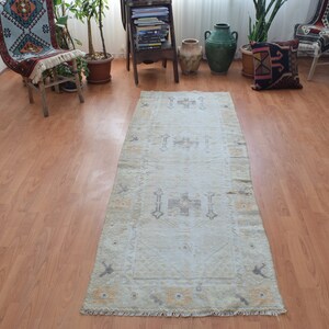 Puede incluir: Una alfombra de pasillo beige claro con un sutil patrón geométrico en un tono beige más oscuro. La alfombra tiene un borde con flecos y está colocada sobre un suelo de madera.