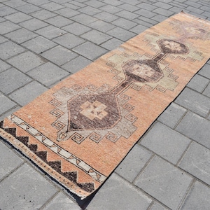 Op de afbeelding: Een lang, rechthoekig tapijt met een perzikkleurige achtergrond en een geometrisch patroon in bruin en wit. Het tapijt heeft franjes aan de uiteinden.