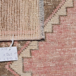 May include: Close-up of a rug's edge with a tag that reads "5058 96 x 242 cm 2'8 x 7'11 ft". The rug is a light pink color with a brown and white pattern.