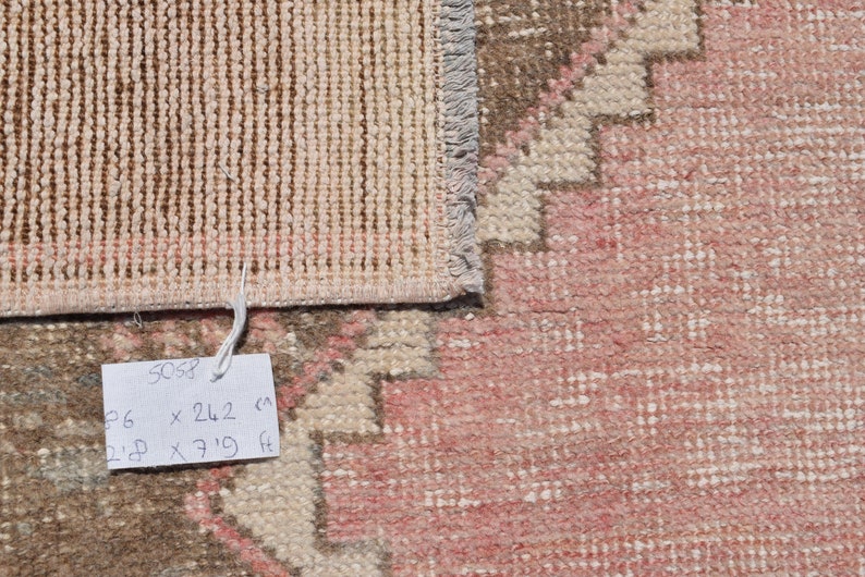 May include: Close-up of a rug's edge with a tag that reads "5058 96 x 242 cm 2'8 x 7'11 ft". The rug is a light pink color with a brown and white pattern.