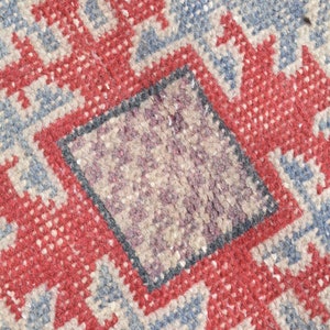 May include: Close-up of a woven rug with a geometric pattern in shades of red, blue, and beige. The rug has a central square with a smaller, darker square inside.
