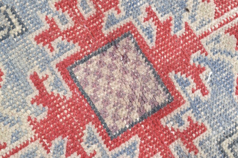 May include: Close-up of a woven rug with a geometric pattern in shades of red, blue, and beige. The rug has a central square with a smaller, darker square inside.