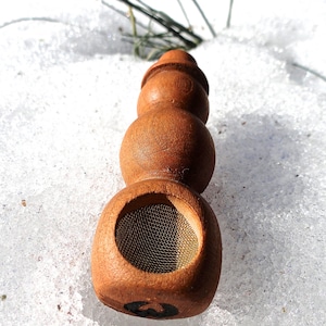 May include: A wooden smoking pipe with a round, bulbous design and a metal screen. The pipe is brown and has a natural wood finish. It is sitting on a white surface.