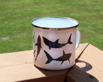 May include: White enamel mug with a black silhouette of sharks on the side. The mug has a silver rim.