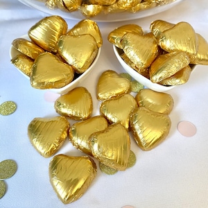 Può includere: Cuori di cioccolato avvolti in foglia d'oro in due ciotole bianche e sparsi su una superficie bianca con coriandoli dorati e rosa.