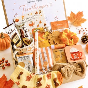 May include: A gift box labelled Welcome to your Treathamper containing a mug, Galaxy hot chocolate, Meredith & Drew biscuits, a candle, orange face mask, eye masks, lip balm, a scrunchie, a headband, and socks with an autumn leaf pattern.