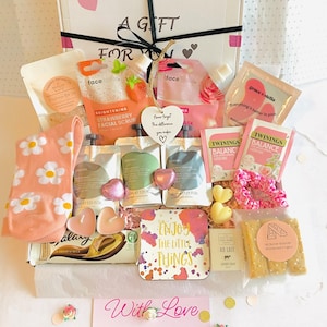 May include: A gift box containing beauty and relaxation products. Items include a strawberry facial scrub, tea bags, soap, and heart-shaped chocolates. The box is decorated with hearts and the phrases "A Gift For You" and "Enjoy the Little Things."