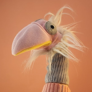 May include: A hand puppet shaped like a bird with a pink fabric head, a yellow inner beak, and a grey ribbed neck. The puppet features a circular grey eye with a black center and long, wispy white faux fur around the neck and head area.