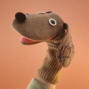 May include: A brown knit dog hand puppet featuring a black pom-pom nose, a white and black circular eye, a pink fabric mouth, and a long floppy ear with a rectangular patch detail. The puppet has a ribbed neck section made of brown fabric.