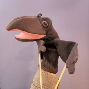 May include: A dark brown bird hand puppet made of knit fabric with a large curved beak, a pink interior mouth, and a small tuft of feathers on the head. The puppet features two extended wings controlled by thin wooden rods attached to the underside.