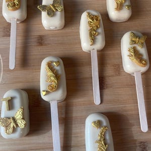 May include: Eight white chocolate-covered popsicles decorated with gold accents. The decorations include butterflies, swirls, and hearts. The popsicles are on a wooden surface.