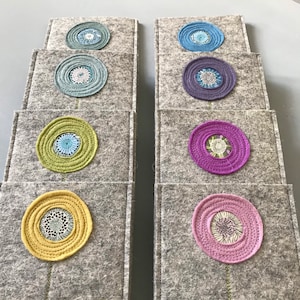 Puede incluir: Un conjunto de 10 posavasos de fieltro con un diseño circular que presenta diferentes colores y patrones. Cada posavasos tiene un solo círculo con un círculo más pequeño en el interior, creando un efecto de capas. Los colores incluyen verde, azul, morado, rosa y amarillo.