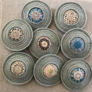 Puede incluir: Ocho posavasos redondos de fieltro con diferentes inserciones de tela estampada. Los posavasos son de color gris azulado con un diseño de círculos concéntricos. Las inserciones de tela presentan varios patrones florales y geométricos en negro, blanco y azul.