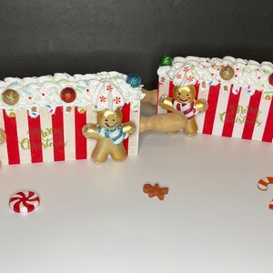 May include: Two decorative rolling pins with red and white stripes, topped with white icing and gingerbread men. The pins have wooden handles and are decorated with sprinkles and baubles. The words "Merry Christmas" are visible on the front.