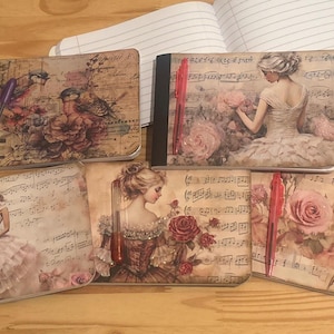 Może przedstawiać: Notatniki z okładkami w stylu vintage z ilustracjami kobiet, róż i nut. Notatniki mają czarne grzbiety i są ułożone na drewnianej powierzchni. Widoczny jest długopis.