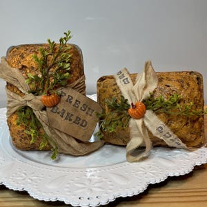 Op de afbeelding: Twee decoratieve broden, gewikkeld in bruin textiel en versierd met groene takjes en oranje pompoenaccenten. Eén brood heeft een label met de tekst "FRESH BAKED". Het brood ligt op een witte, decoratieve schaal.