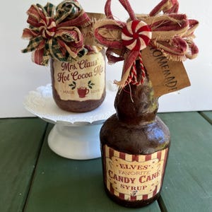 May include: Two decorative jars with festive holiday themes. One jar is labeled "Mrs. Claus Hot Cocoa Mix" and the other is labeled "Elves' Favorite Candy Cane Syrup." Both jars are adorned with bows and ribbons, perfect for holiday decor.