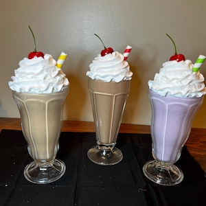 Puede incluir: Tres vasos decorativos de batidos. Cada vaso contiene un líquido de diferente color: beige, marrón y lavanda. Cada batido está cubierto con crema batida, una cereza y una pajita. Las pajitas son amarillas, rojas y blancas, y verdes y blancas.