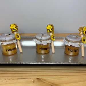 Puede incluir: Tres pequeños tarros de miel con tapas decorativas y cucharas de miel de madera. Cada tarro tiene una etiqueta que dice "Honey". Las cucharas de miel están rematadas con un diseño de abeja amarilla y girasol. Los tarros están sobre una bandeja plateada.